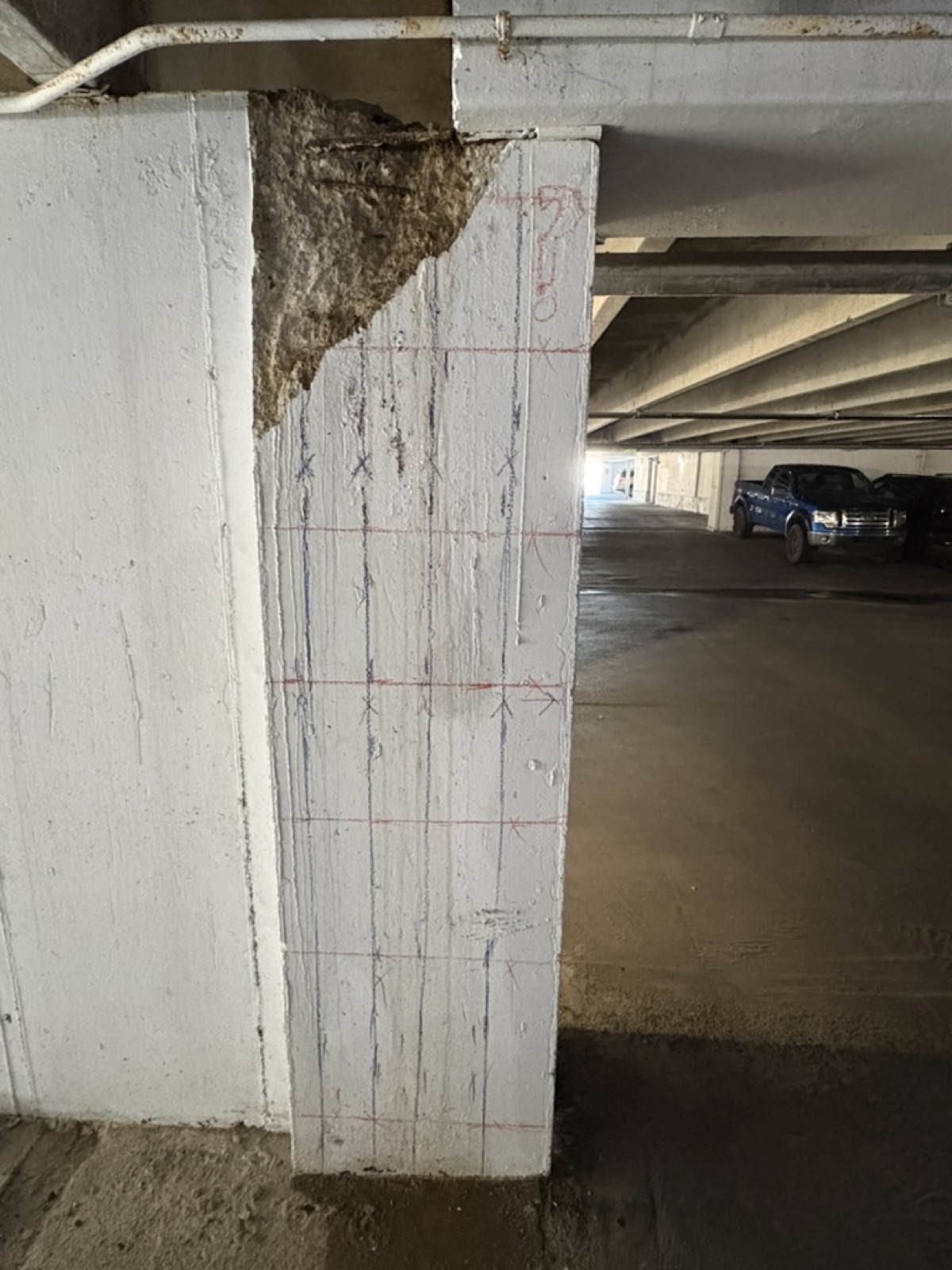 Close view of a marked column face in a parking garage showing bar lines and X marks, with spalled concrete exposing aggregate at the top corner