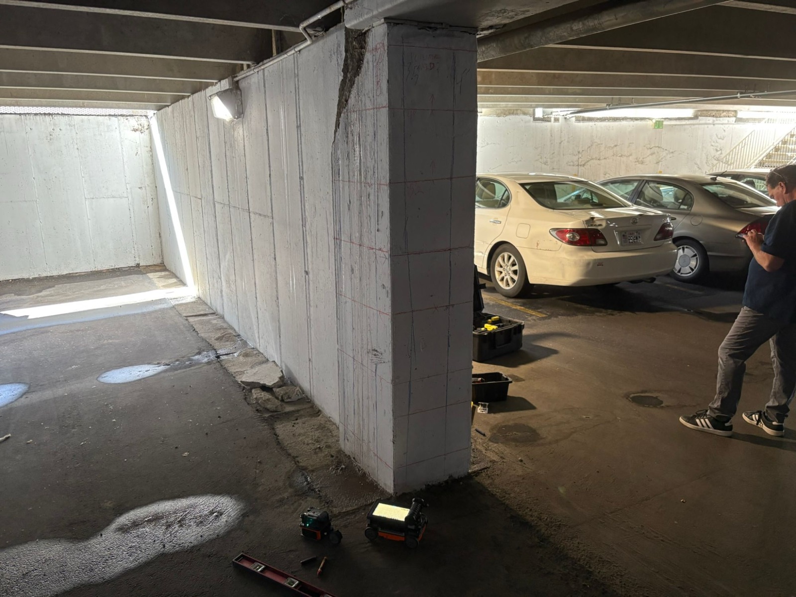 Long concrete shear wall in a parking garage marked over its full height, radar units on the floor, parked cars beyond